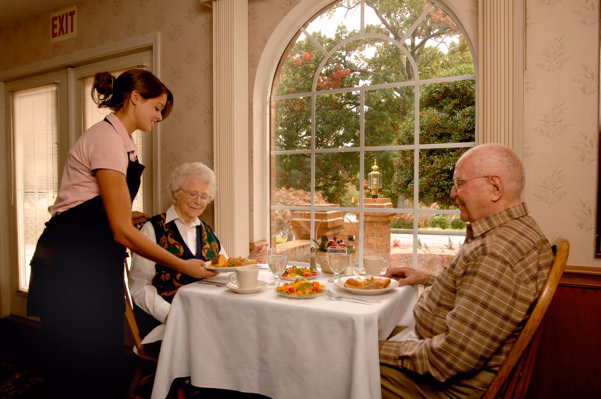 A waitress serving food to an elderly woman seated at a table with an elderly man in a dining room with a large arched window showing trees and a brick exterior.