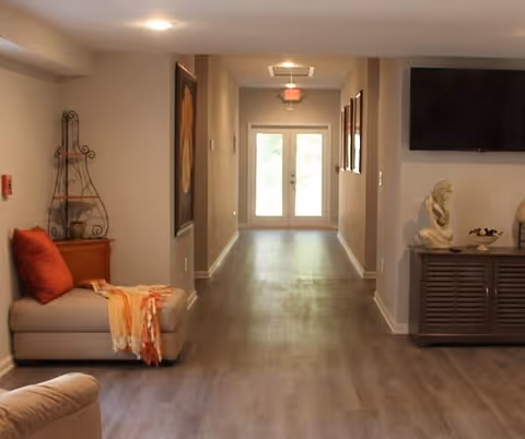 A well-lit hallway in a senior living facility with light-colored walls and wood flooring. On the left side, there is a beige cushioned bench with a red pillow and an orange throw blanket. On the right side, there is a cabinet with decorative items including a sculpture and a bowl, and a wall-mounted flat-screen TV above it. At the end of the hallway, double glass doors allow natural light to enter.