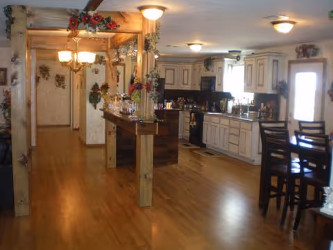 Interior view of a kitchen and dining area with wooden floors and white cabinets. The kitchen features a stove, sink, and countertop with various items on it. There is a wooden dining table with chairs on the right side and decorative plants and flowers around the room. The space is well-lit with ceiling lights and a chandelier near a wooden beam structure.