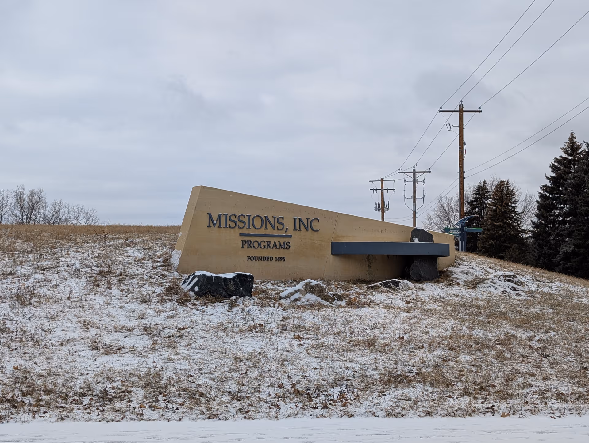 A large beige stone sign on a snowy grassy hill with the text 'MISSIONS, INC PROGRAMS FOUNDED 1895'. There are some rocks around the base of the sign and utility poles and trees in the background under a cloudy sky.