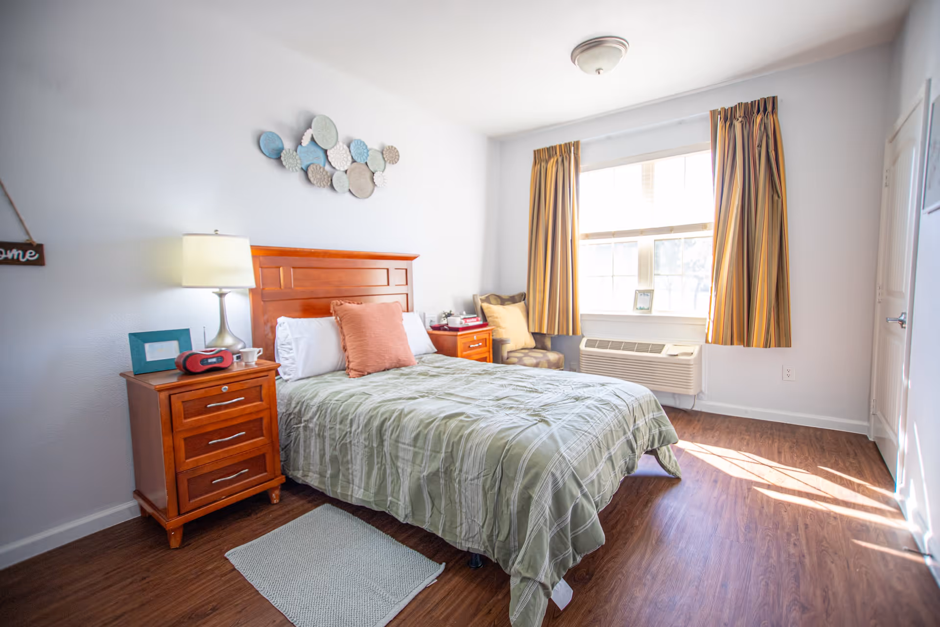 A bright bedroom with a wooden bed frame and green striped bedding, two wooden nightstands with a lamp and alarm clock, a cushioned chair near a window with striped curtains, and wooden flooring.