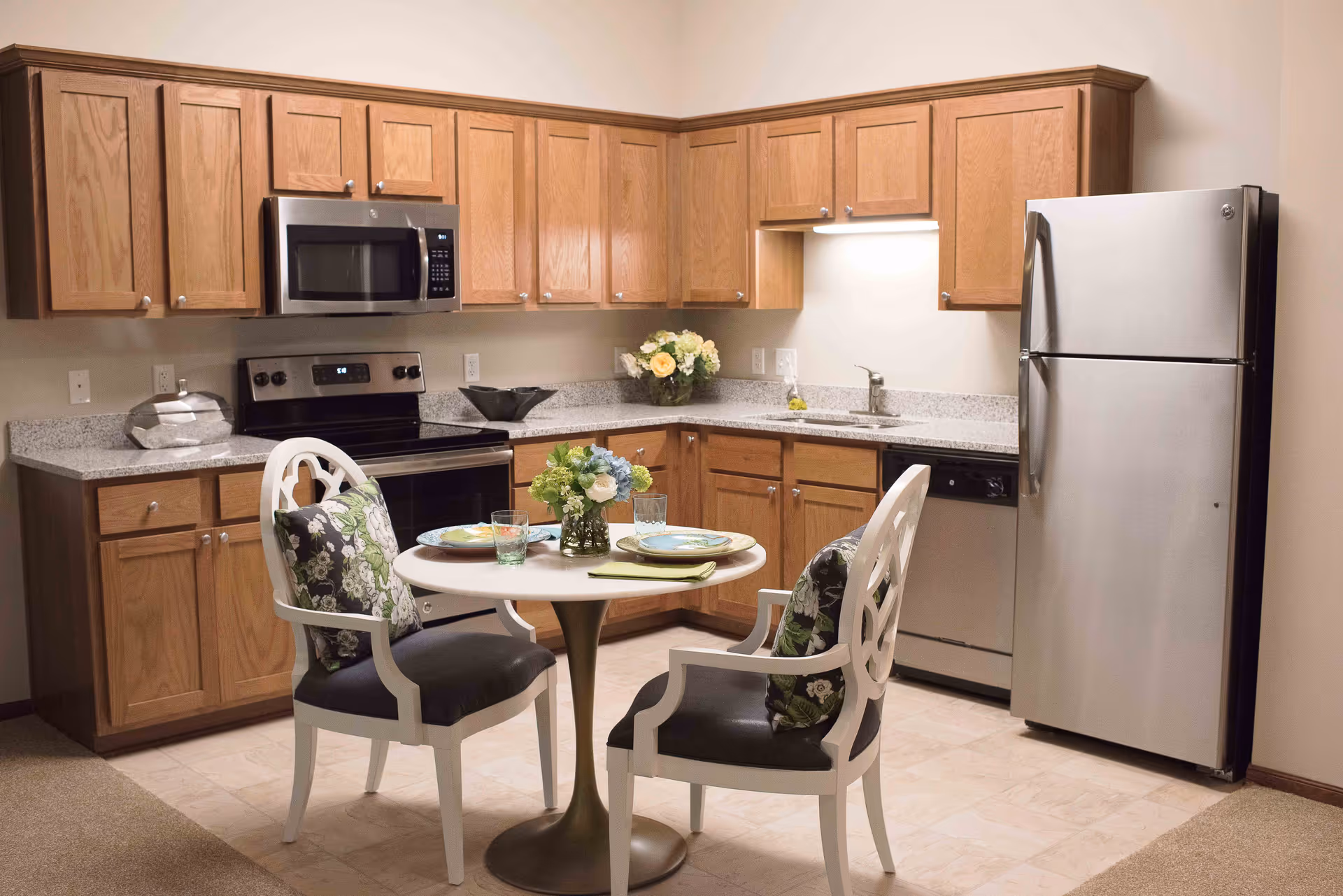 A kitchen area with wooden cabinets, a stainless steel refrigerator, microwave, oven, and dishwasher. There is a small round dining table with two chairs, each with a floral cushion. The table is set with plates, glasses, and a small flower arrangement.