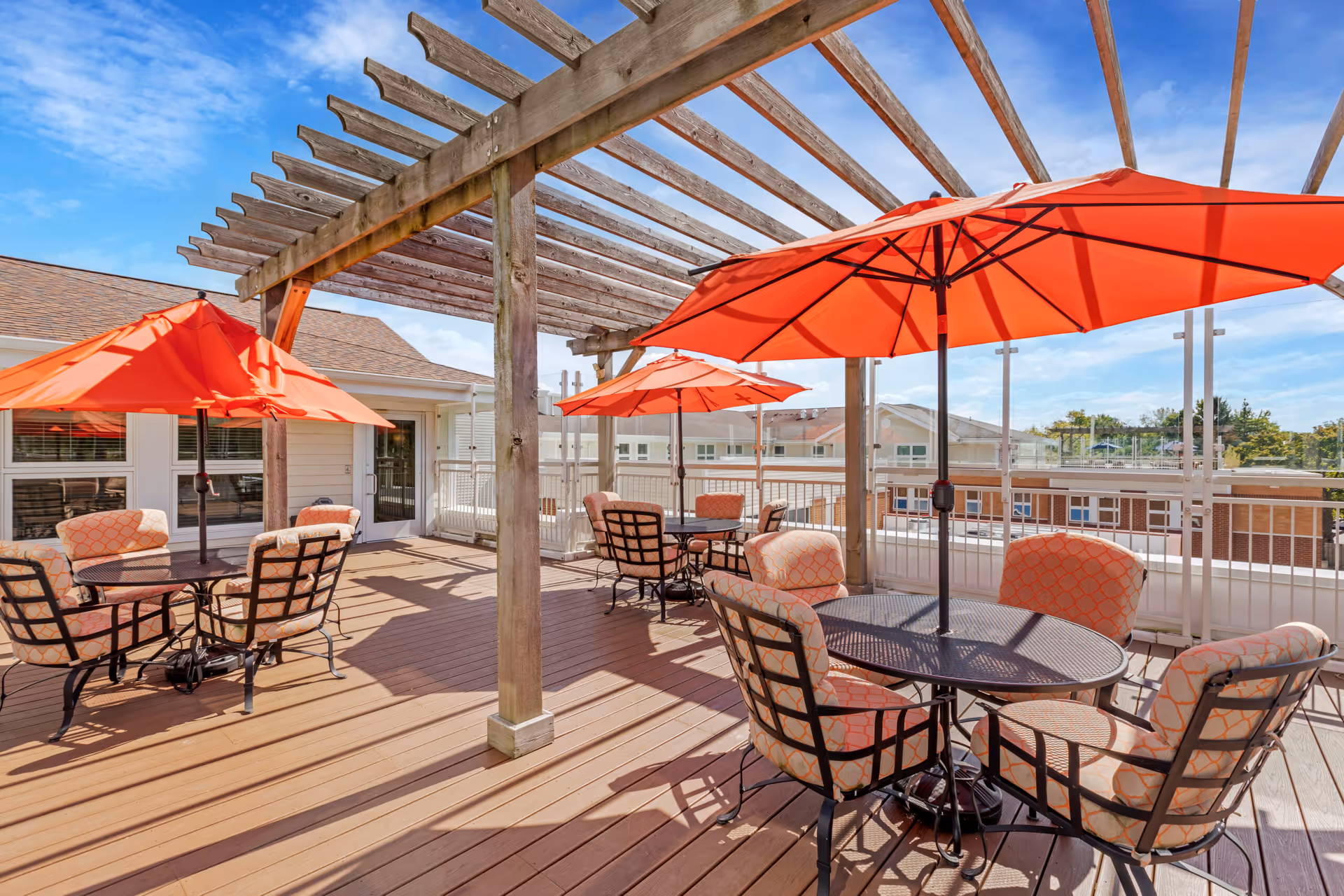 Outdoor patio area with wooden deck flooring, several round tables with cushioned chairs, and bright orange umbrellas providing shade. A wooden pergola structure extends overhead, and buildings are visible in the background under a clear blue sky.