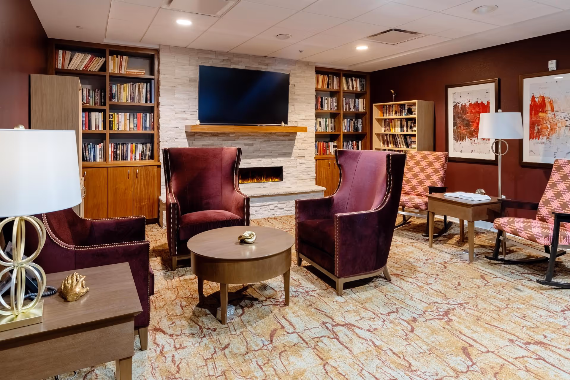 A cozy living room with four purple armchairs arranged around a round wooden coffee table. Behind the chairs is a modern electric fireplace set in a white stone wall with a flat-screen TV mounted above it. On either side of the fireplace are built-in wooden bookshelves filled with books. The room has warm brown walls adorned with two abstract paintings and is softly lit by table lamps and a floor lamp. The carpet has a beige and brown pattern.