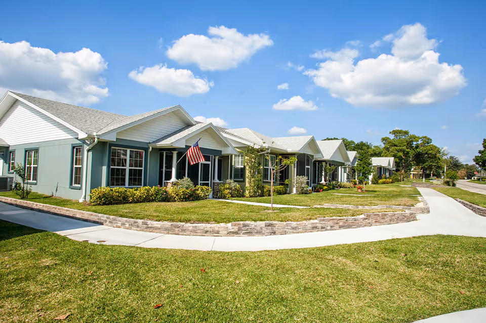Exterior view of a single-story residential building with multiple units, featuring green and white walls, large windows, and a front porch with an American flag. The building is surrounded by well-maintained grass, small trees, and shrubs, with a curved concrete walkway in front under a blue sky with scattered clouds.
