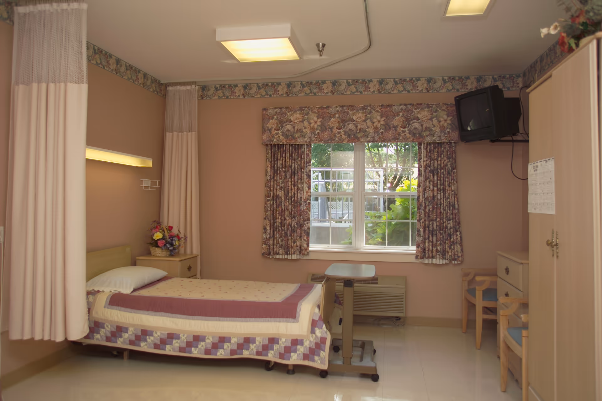 A nursing home bedroom with a single bed covered with a quilted blanket, a bedside table with a flower arrangement, floral curtains on the window, a small table on wheels, a wall-mounted TV, two chairs, and a wardrobe.