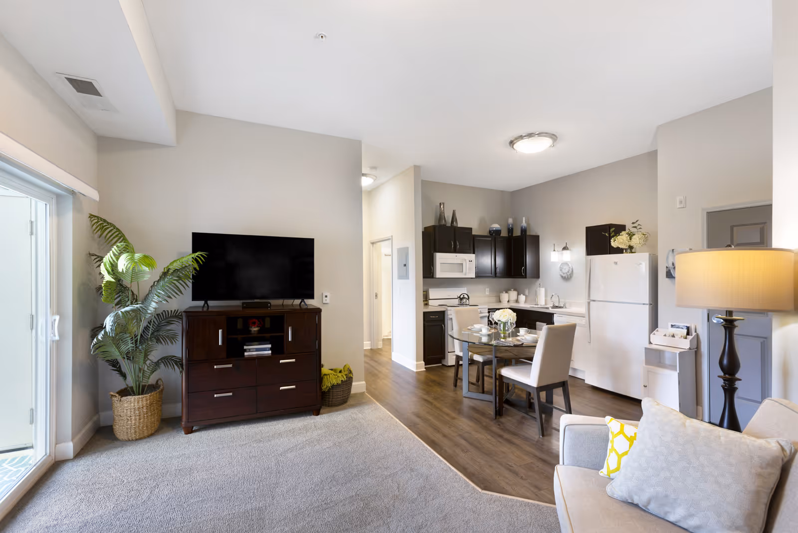 A bright and modern living area with a beige sofa and yellow-accented pillows, a dark wooden TV stand with a flat-screen TV, a potted plant next to sliding glass doors, and an open kitchen with dark cabinets, white appliances, and a glass dining table with four chairs set for a meal.