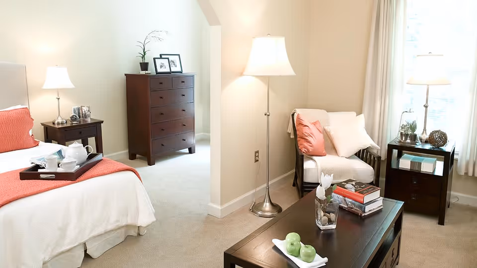 A bright and cozy bedroom with a white bed featuring coral-colored pillows and a matching throw blanket. A dark wooden nightstand with a lamp and a tray with a teapot and cups is beside the bed. In the background, there is a dark wooden dresser with framed photos and a small plant. A sitting area with a cushioned chair, coral and white pillows, a floor lamp, and a dark wooden coffee table with books and decorative items is also visible. Light streams in through a window with white curtains.