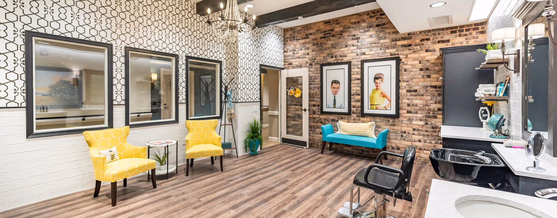 A stylish interior room with a modern design featuring patterned wallpaper, a brick accent wall, and wood flooring. The room includes two yellow armchairs with a small round table between them, a teal bench with a cushion, three framed portraits on the brick wall, and a black salon chair in front of a black sink and countertop with shelves holding towels and hair products.