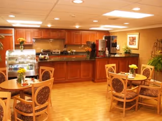 A warm and inviting dining area with wooden tables and chairs, each table decorated with a small vase of yellow flowers. In the background, there is a kitchen counter with wooden cabinets, a coffee machine, and a display case with food items. The room has bright ceiling lights and a framed picture on the wall.