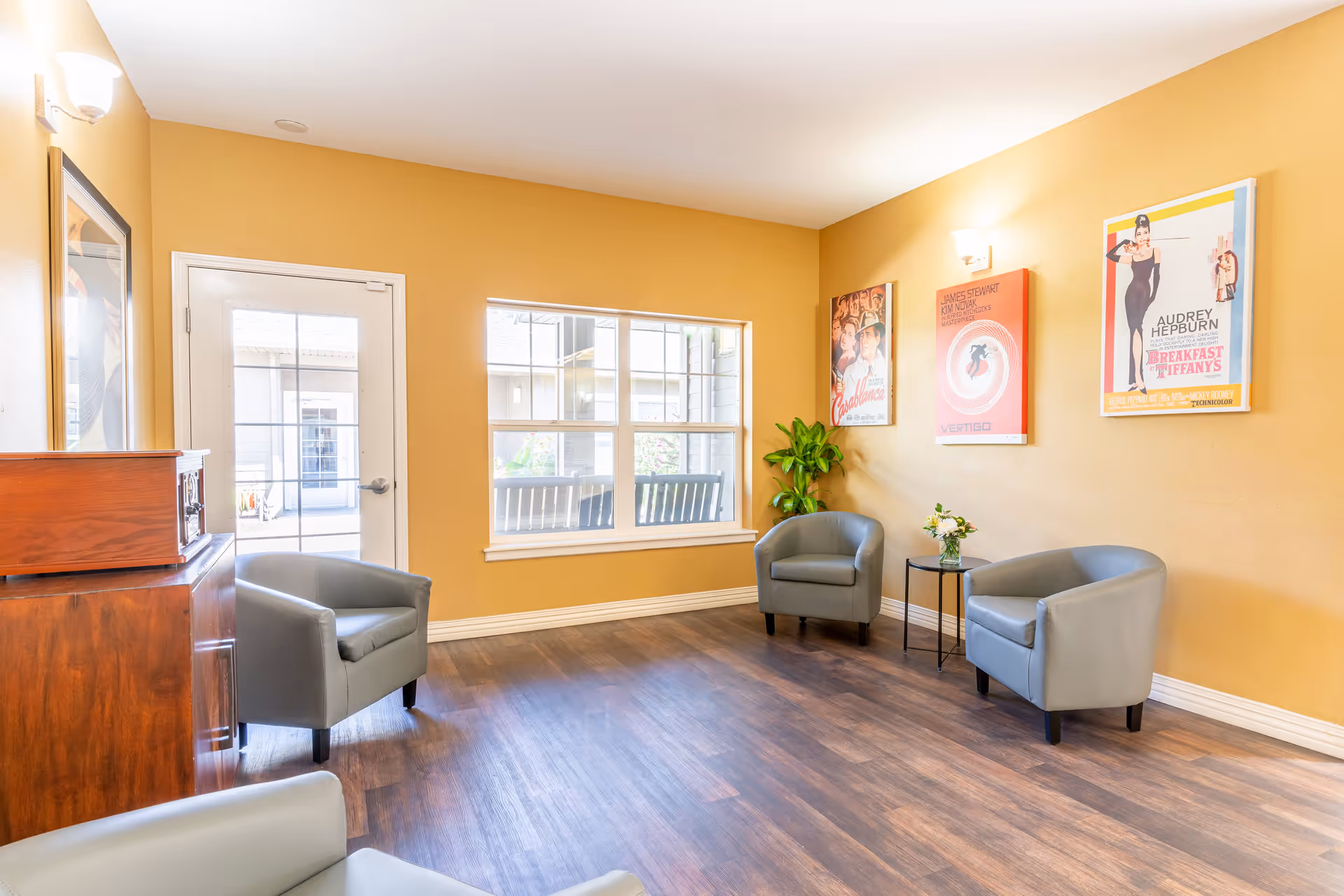 A cozy sitting area with three gray armchairs arranged around a small black side table with a flower vase. The walls are painted a warm yellow and decorated with three vintage movie posters. There is a window and a glass door letting in natural light, and a wooden cabinet is visible on the left side. The floor is dark wood.