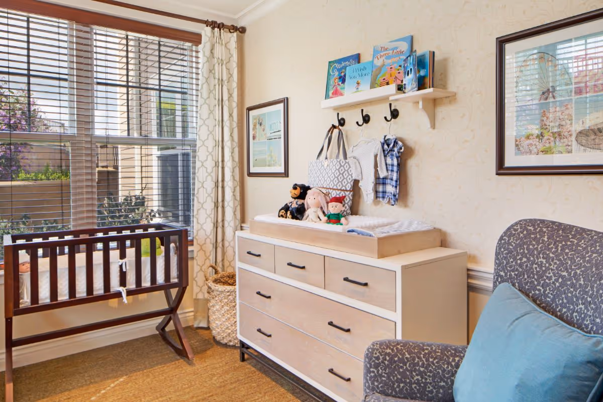 A cozy nursery room with a wooden crib near a large window with blinds and patterned curtains. A dresser with a changing pad on top is against the wall, decorated with stuffed animals and children's clothes hanging on hooks above. Two framed pictures hang on the wallpapered wall, and a patterned armchair with a blue cushion is partially visible.