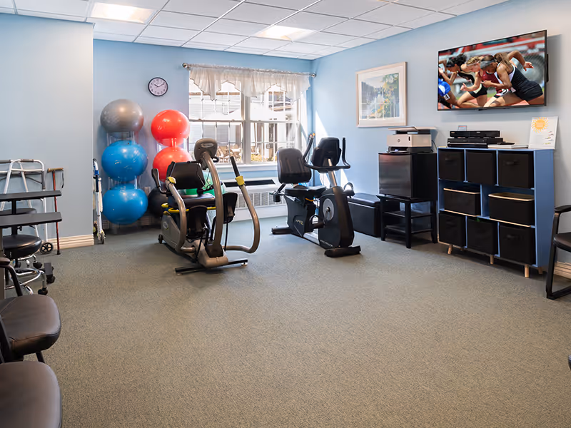 Bright exercise room with recumbent exercise bikes, stacked stability balls, storage cubbies and a wall-mounted TV.