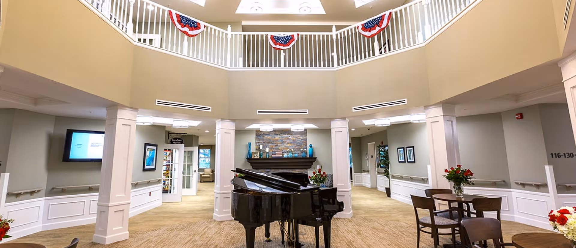 Interior view of a senior living facility lobby area with a black grand piano in the center, surrounded by round tables and chairs with flower vases. The space features beige walls, white pillars, a stone fireplace, and patriotic red, white, and blue bunting hanging from the upper balcony railing.