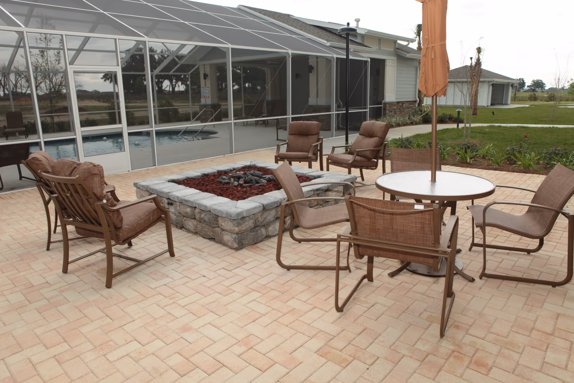 Outdoor patio area with cushioned chairs arranged around a stone fire pit and a round table with an umbrella. In the background, there is a screened-in pool area and a building with a garage. The patio is paved with light-colored bricks and surrounded by grass and landscaping.