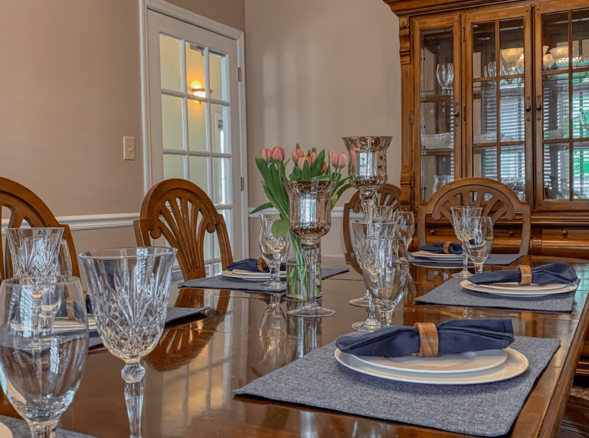 A formal dining room with a polished wooden table set with plates, blue napkins in wooden rings, crystal glassware, and silver candle holders. A vase with pink tulips is placed in the center of the table. Wooden chairs surround the table, and a wooden china cabinet with glass doors displaying glassware is visible in the background. A white door with glass panels is on the left wall.
