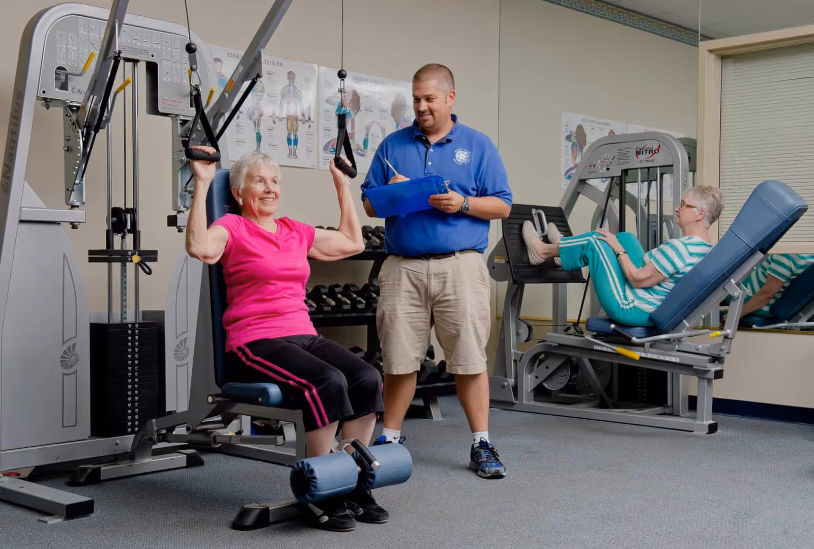 Two elderly women exercising on gym machines in a fitness room while a male trainer in a blue polo shirt takes notes on a clipboard.