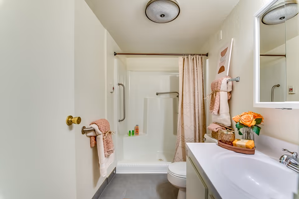 A clean and well-lit bathroom featuring a white shower with a beige patterned curtain, a white toilet, and a white sink countertop. The bathroom has light-colored walls and gray floor tiles. Towels in shades of pink and white are neatly hung on towel racks, and a small vase with orange roses is placed on the countertop next to a soap dish and other toiletries. A round ceiling light fixture is visible above.