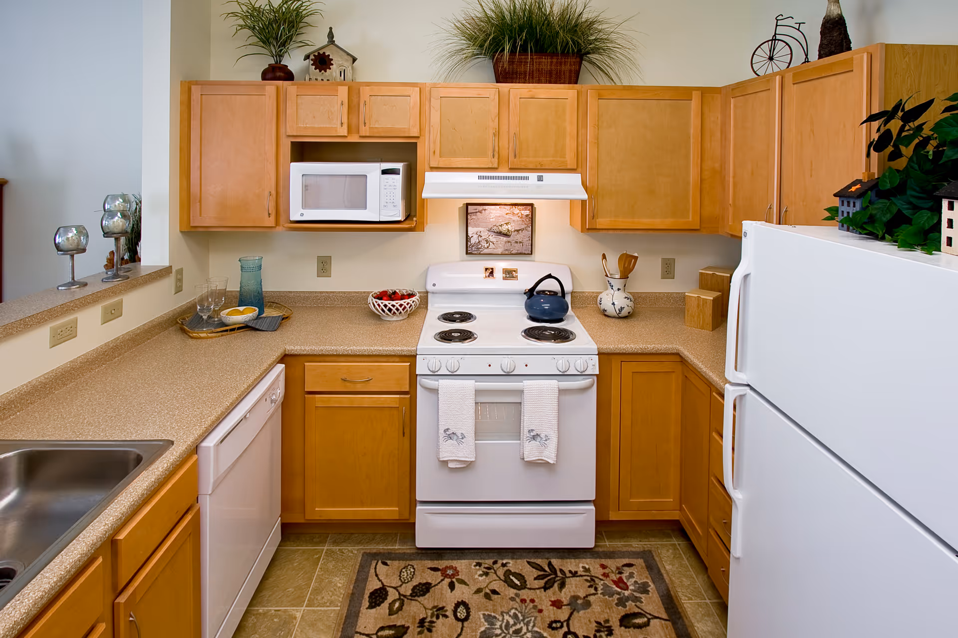 Compact kitchen with light wood cabinets, white stove and refrigerator, microwave, sink, and countertop decor.