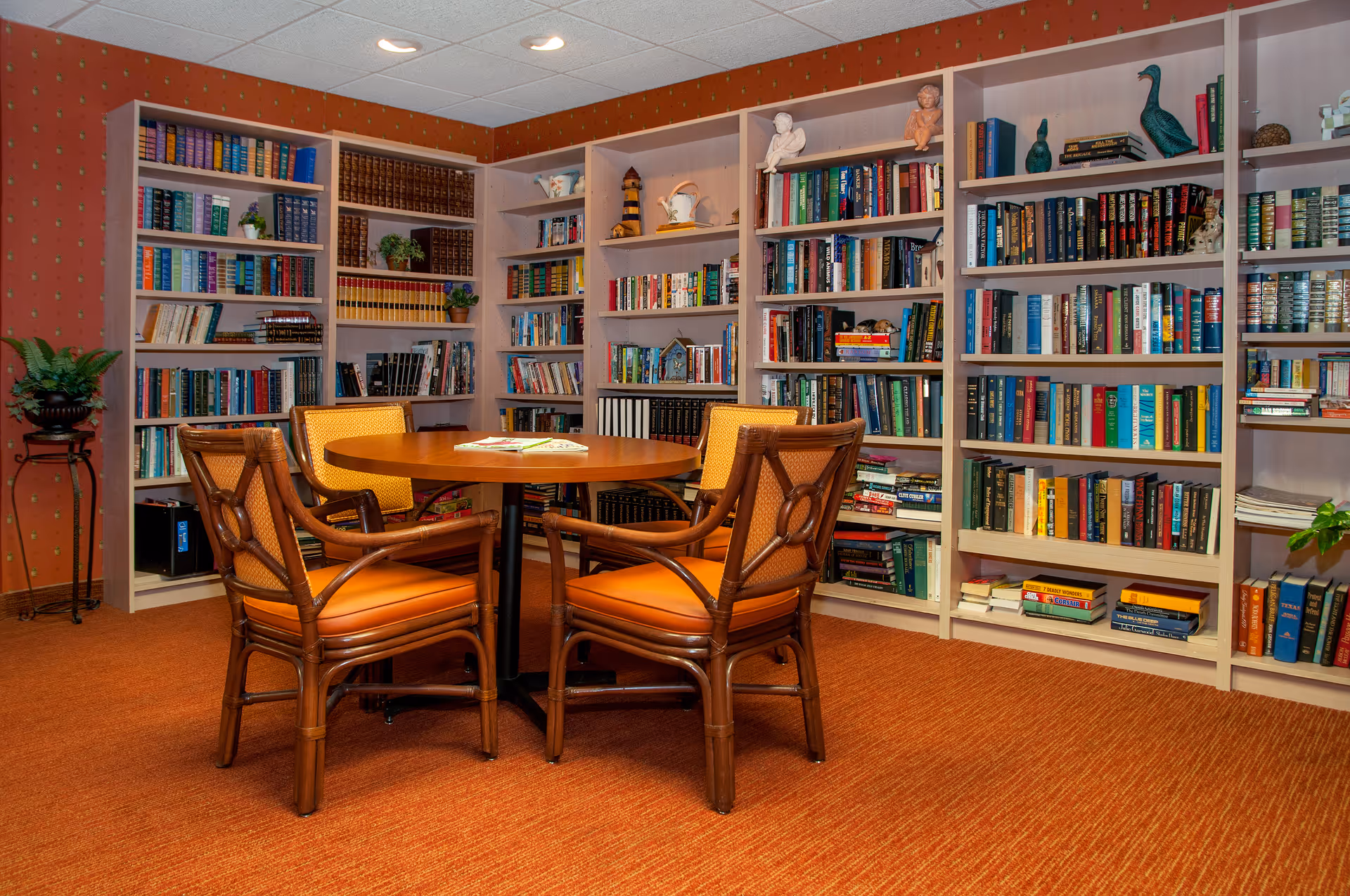 A cozy library room with multiple white bookshelves filled with books and decorative items. In the center, there is a round wooden table surrounded by four wooden chairs with orange cushions. The room has orange carpeting and warm lighting.