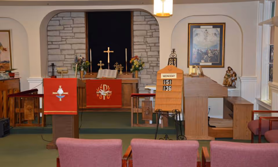 Interior view of a small chapel or worship room with a stone wall altar featuring a cross, candles, flowers, and religious symbols. There are red cloths with Christian symbols draped over wooden podiums, a wooden lectern, and a hymn board displaying numbers. Chairs with red upholstery are visible in the foreground, and religious artwork and statues are on the walls and shelves.