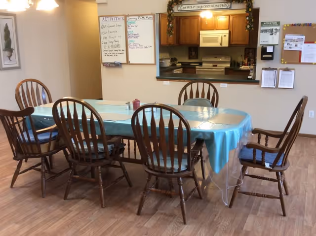 A dining area with a rectangular table covered with a blue tablecloth and six wooden chairs with blue cushions. In the background, there is a kitchen pass-through window with wooden cabinets, a microwave, and a stove. On the wall beside the kitchen window, there are bulletin boards, a whiteboard with activities listed, and some decorative greenery above the kitchen window.
