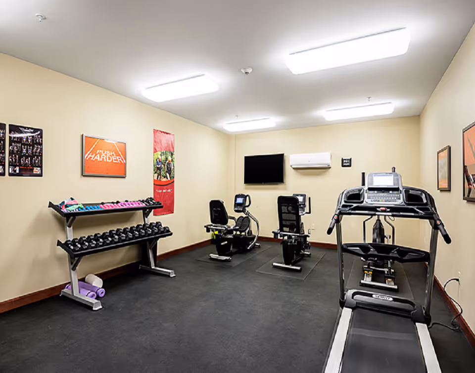 A small fitness room with exercise equipment including a treadmill, two recumbent bikes, and a rack of dumbbells and hand weights. The room has beige walls with motivational posters, a wall-mounted TV, and bright overhead lighting.
