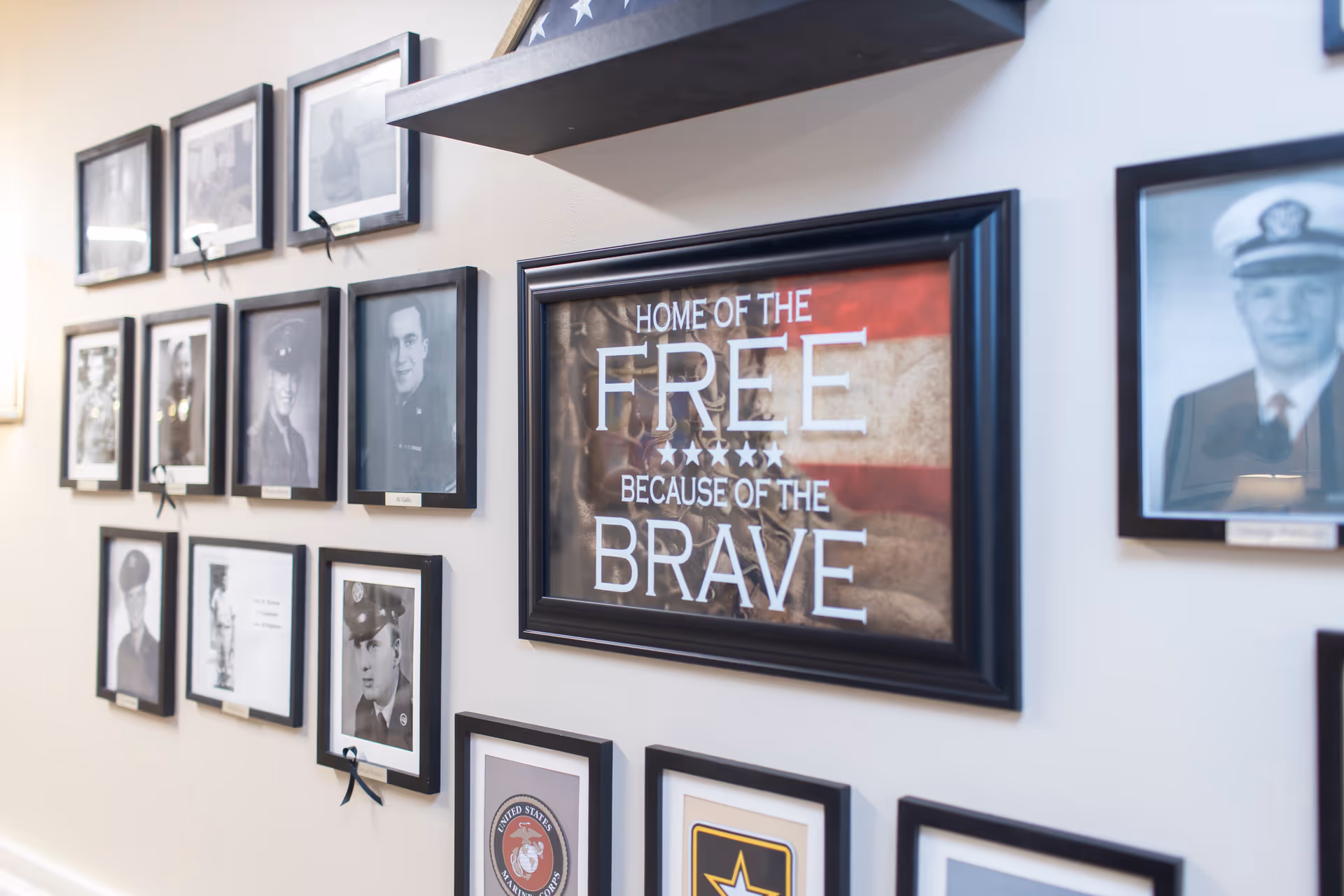 A wall display featuring framed black and white photographs of military personnel and patriotic symbols, including a central framed sign with an American flag background that reads 'Home of the Free Because of the Brave.'