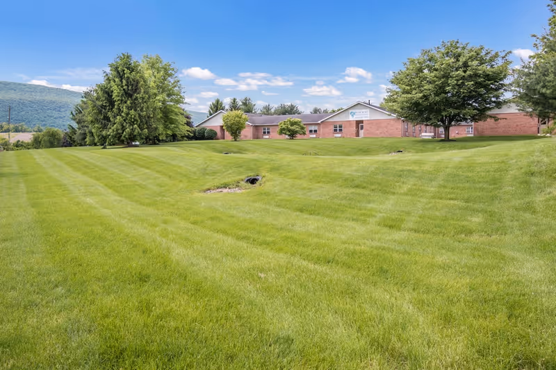 Wide view of a large, well-maintained grassy lawn with a few trees and a brick building in the background under a blue sky with scattered clouds. The building has a sign that reads 'Celebration Villa of Reedsville'.