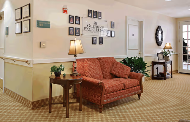 A cozy seating area in a senior living facility hallway featuring a red patterned loveseat with matching cushions, a wooden side table with a lamp and a small plant, framed photos and a 'Gallery of Excellence' sign on the wall, another table with a lamp and decorative items, a round mirror, and a door leading outside.