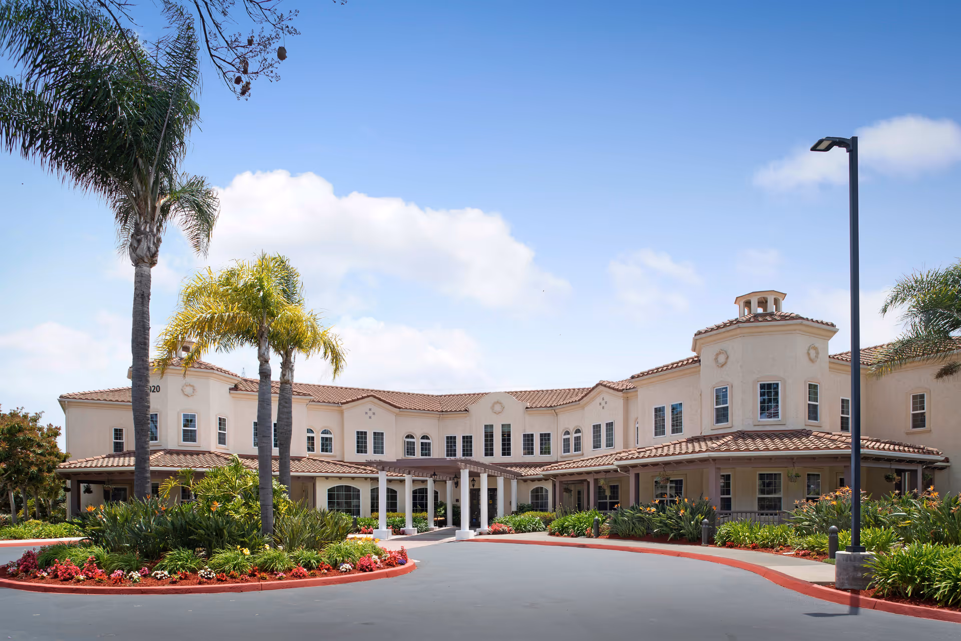 Exterior view of a two-story senior living facility building with beige walls and a red-tiled roof, surrounded by palm trees and landscaped greenery under a blue sky with some clouds.