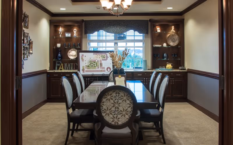 A formal dining room with a long rectangular table surrounded by eight upholstered chairs. The room features built-in dark wood cabinets and shelves on the back wall, decorated with various items including a framed picture, vases, and a decorative plate. A window with blinds and a valance is centered on the back wall, allowing natural light into the room. A chandelier hangs from the ceiling above the table.