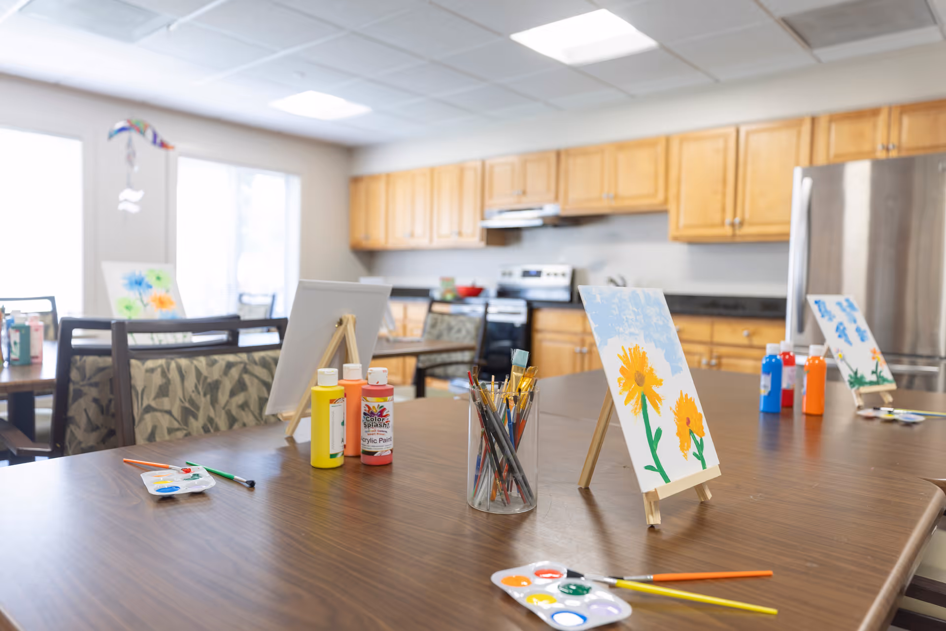 A bright room with wooden cabinets and a stainless steel refrigerator in the background, featuring tables set up for painting activities with canvases on easels, paint bottles, brushes, and palettes with paint.
