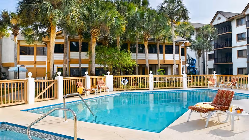 Outdoor swimming pool area at Vitality Living Baypoint Village with lounge chairs and palm trees surrounding the pool. The pool is fenced with a wooden railing, and there are multi-story buildings in the background.