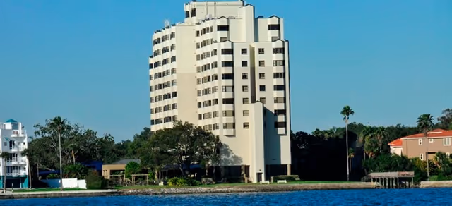 A tall, multi-story white residential building with balconies, situated near a body of water with trees and other smaller buildings nearby under a clear blue sky.