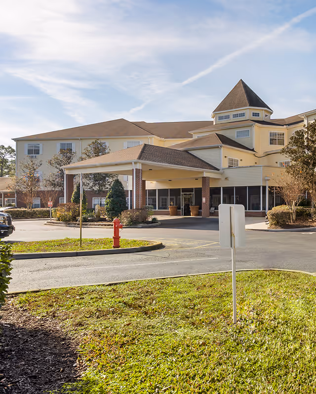 Exterior view of Brookdale Lake Mary senior living facility showing the main entrance with a covered drop-off area, surrounded by landscaped greenery and a parking lot under a partly cloudy sky.