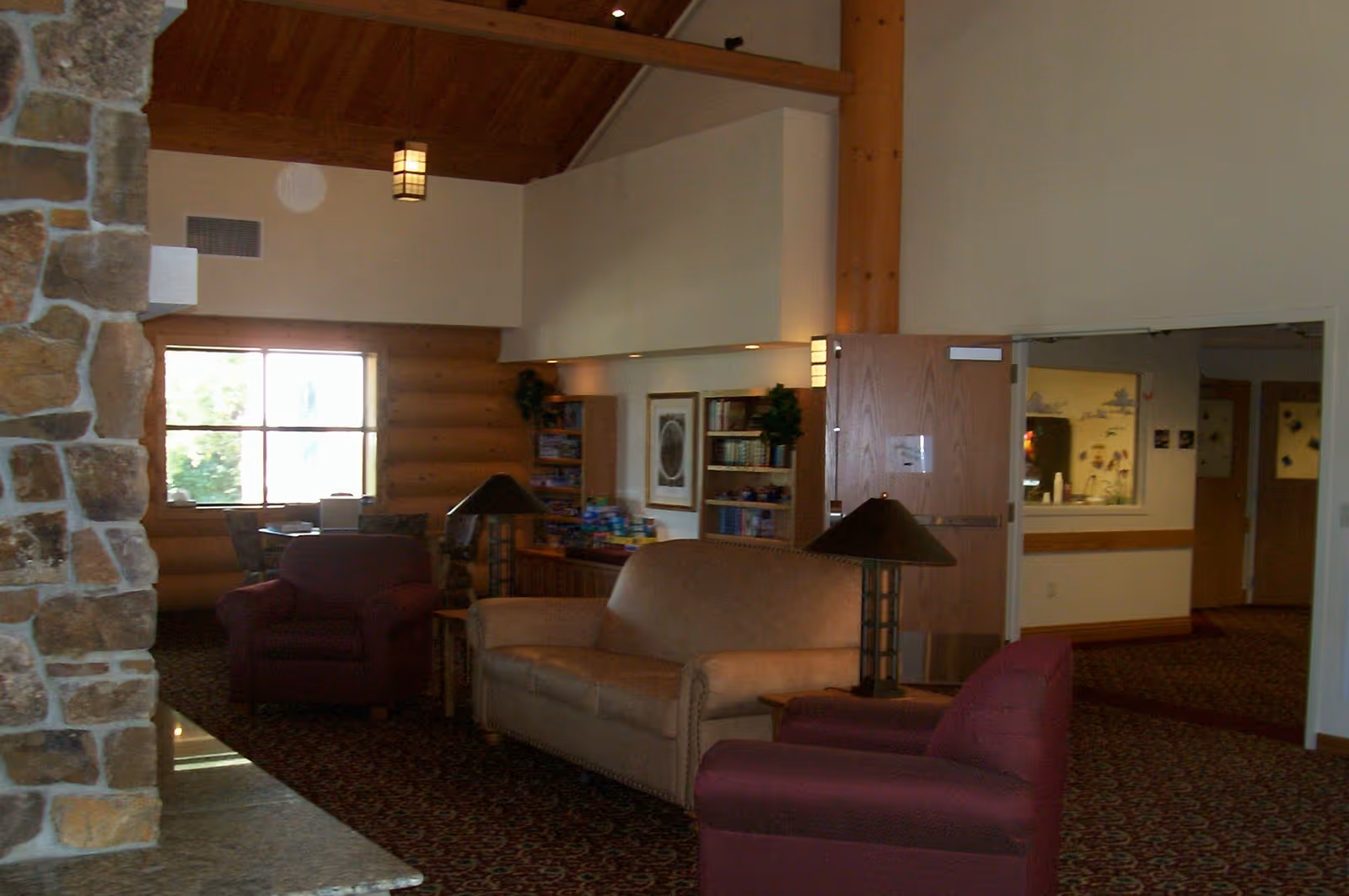 Interior view of a cozy living room area in an assisted living facility featuring a beige sofa, two maroon armchairs, a stone fireplace on the left, wooden ceiling beams, carpeted floor, table lamps, bookshelves, and a window letting in natural light.