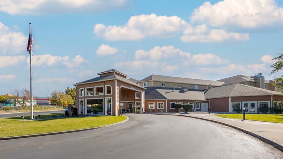 Exterior view of Brookdale Richland senior living facility showing a curved driveway leading to the main entrance with a covered drop-off area, an American flag on a flagpole, green lawns, and a partly cloudy sky.