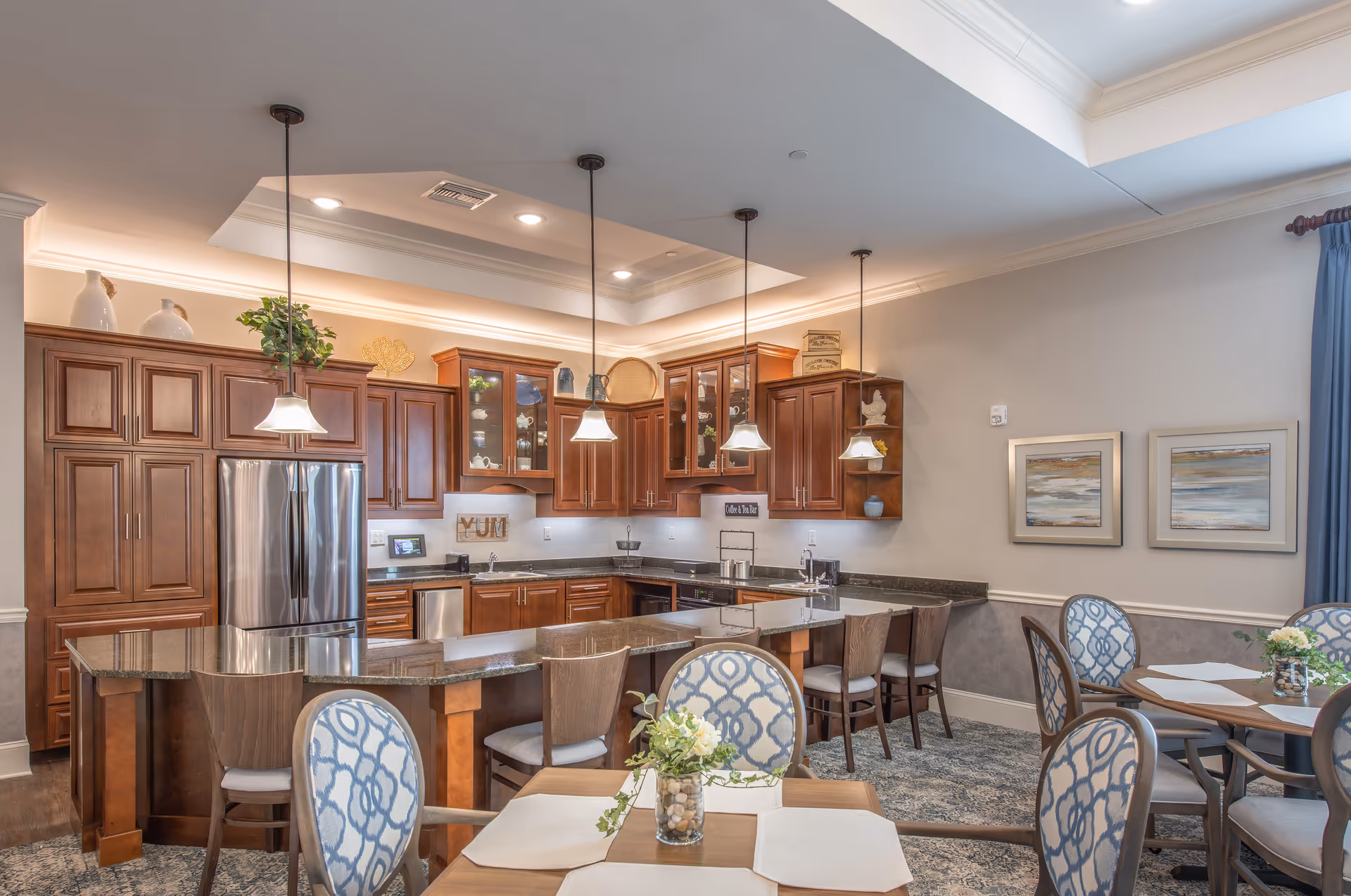 Bright communal kitchen and dining area with dark wood cabinetry, a large island with bar stools, pendant lights, and several dining tables.