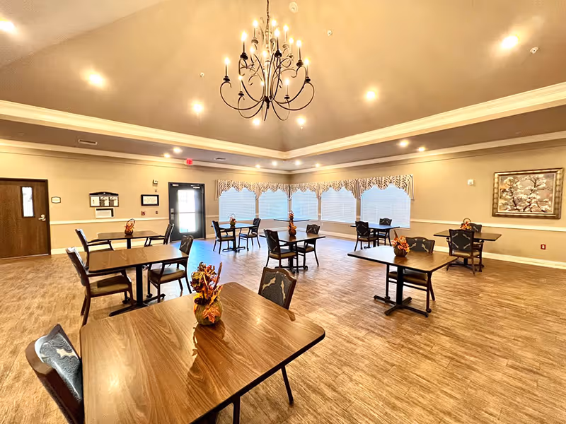A spacious dining room with several wooden tables and chairs arranged neatly. Each table has a small autumn-themed centerpiece. The room features large windows with patterned valances, a chandelier hanging from the ceiling, recessed lighting, and light-colored walls with a framed artwork. The floor has a wood-like finish.