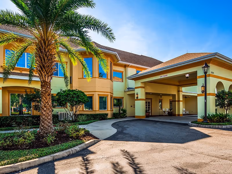 Exterior view of The Windsor at Ortega senior living facility showing a two-story building with yellow and beige walls, large windows, a covered entrance with a driveway, palm trees, and landscaped greenery under a clear blue sky.