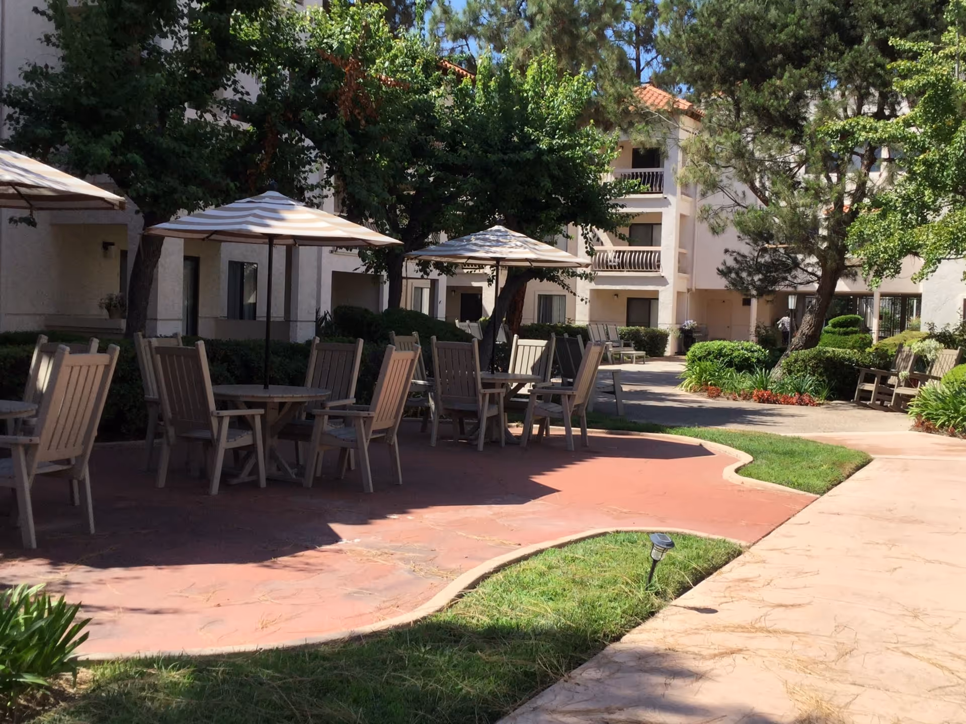 Sunlit courtyard with patio tables, umbrellas, and chairs in front of a multi-story senior living building.