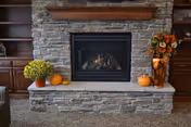 A stone fireplace with a wooden mantel in a cozy living room setting. The mantel is decorated with a vase of autumn flowers and small pumpkins. Built-in wooden shelves flank the fireplace on both sides.