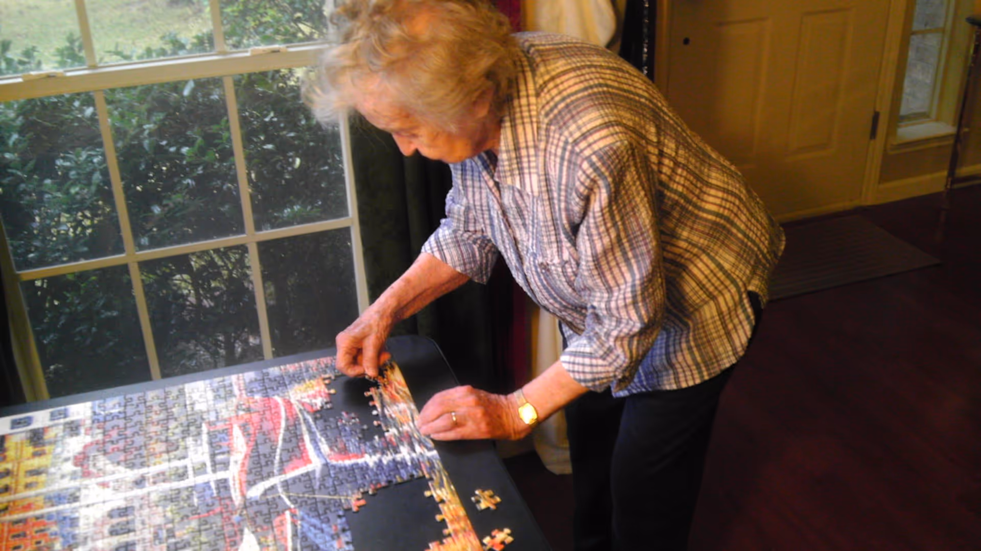 An older woman working on a jigsaw puzzle on a table beside a window in a living room.