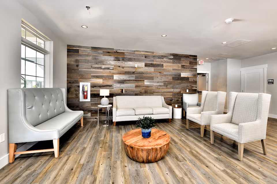 A modern and bright seating area with a wooden accent wall, light-colored upholstered chairs and sofas, a round wooden coffee table with a small potted plant, and a window letting in natural light.