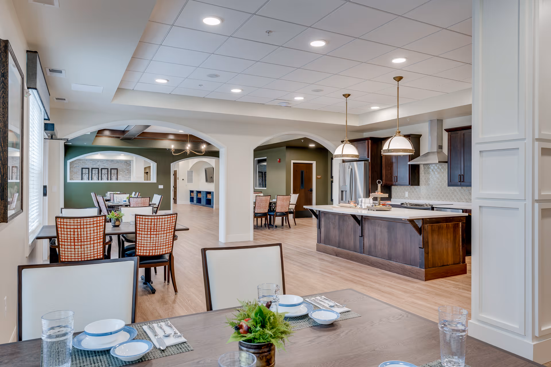 A spacious and well-lit dining area in a senior living facility featuring multiple tables with chairs, some set with plates, bowls, and glasses. The room has light wood flooring, white walls, and a ceiling with recessed lighting. In the background, there is a modern kitchen with dark wood cabinetry, a large island with pendant lights, stainless steel appliances, and a range hood. The dining area extends into another seating space with additional tables and chairs, separated by white archways.