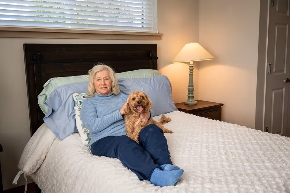An elderly woman with gray hair wearing a light blue sweater and dark blue pants is sitting on a bed with a white textured blanket, holding a small brown dog. Behind her is a dark wooden headboard, pillows, and a nightstand with a lamp. A window with blinds is above the bed.