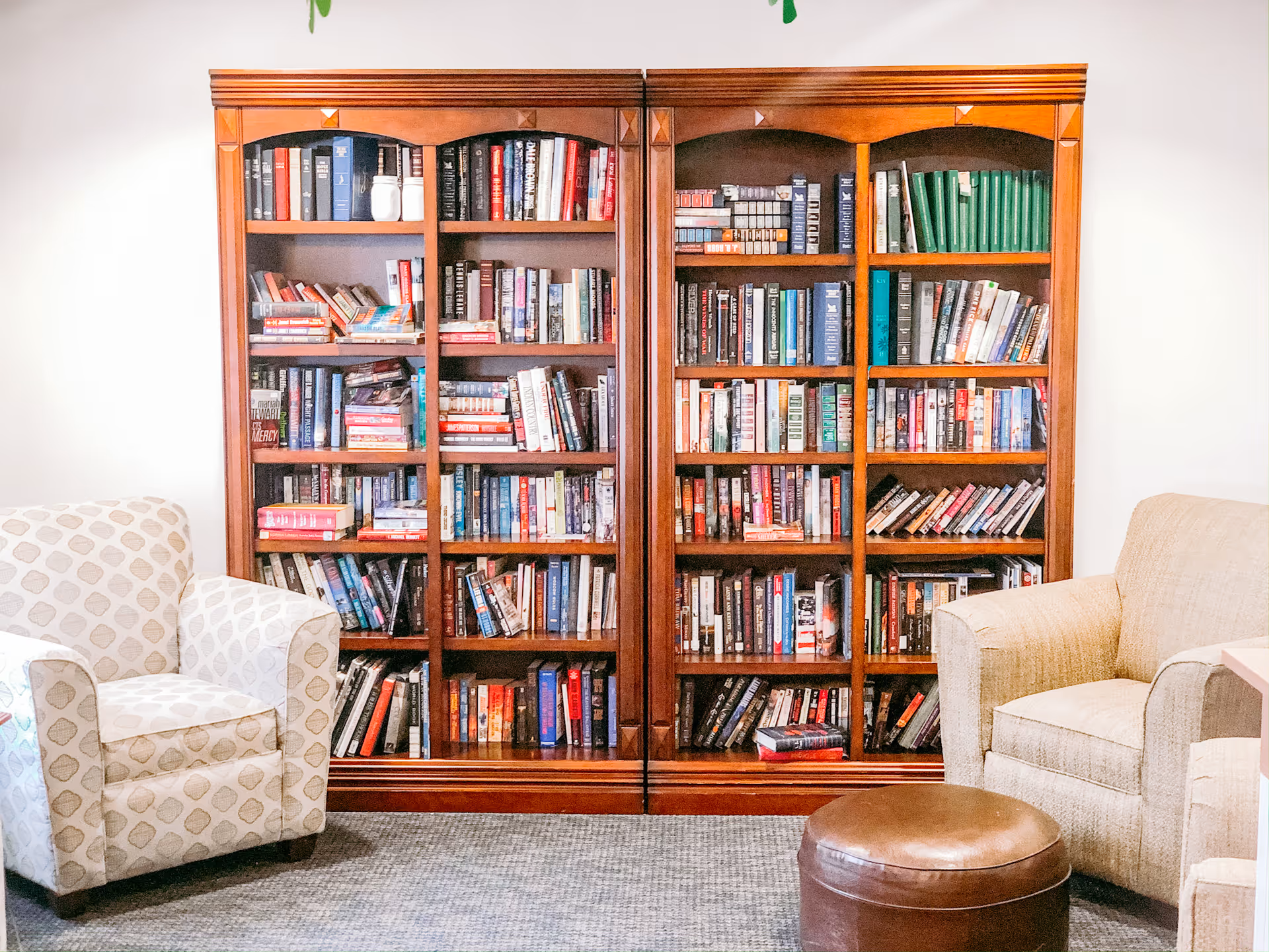 A cozy reading area in a senior living facility featuring two upholstered armchairs, a round brown leather ottoman, and a large wooden bookshelf filled with various books against a white wall.