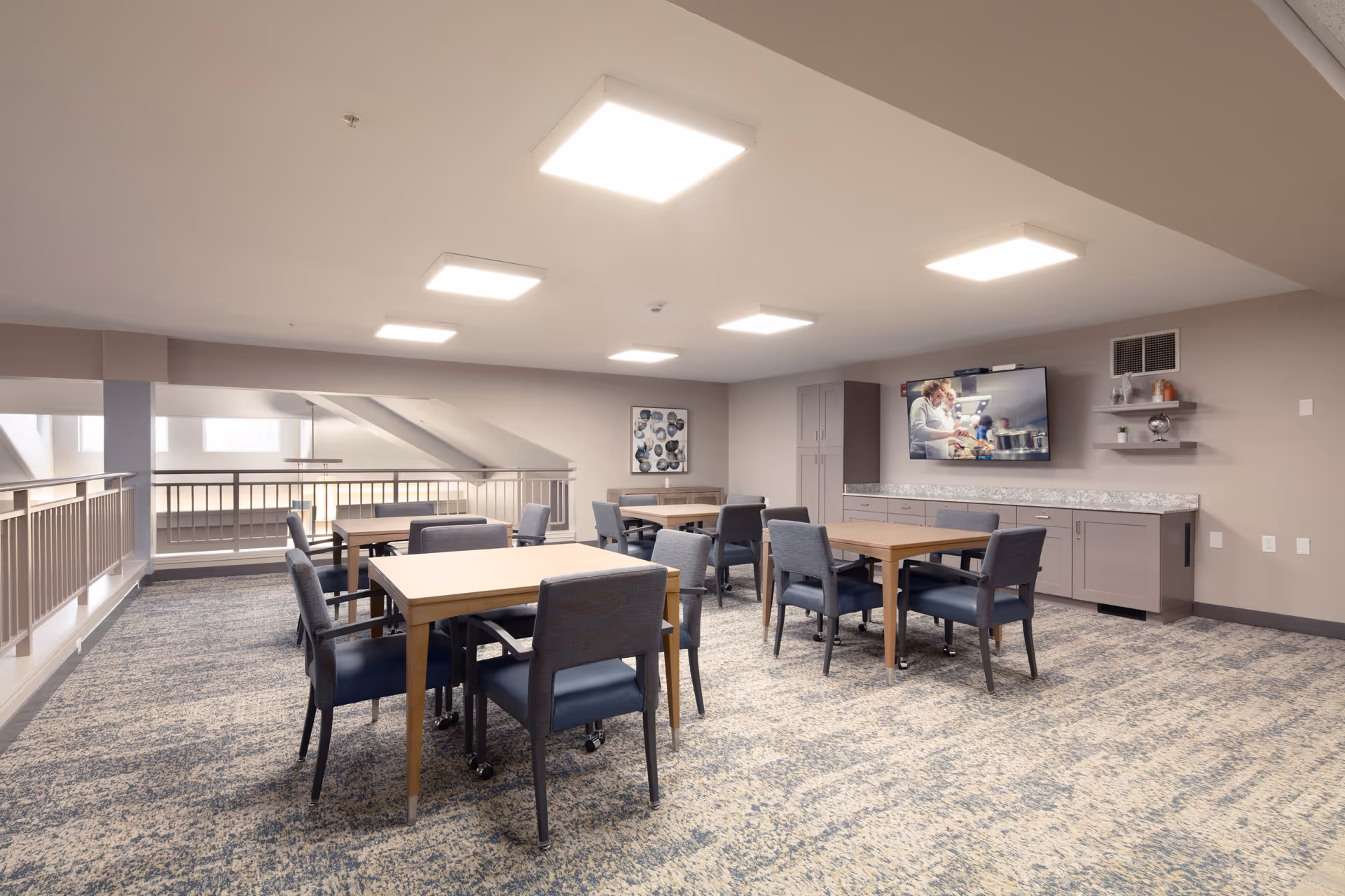 A well-lit common area with several wooden tables and cushioned chairs arranged on a patterned carpet. The room has a neutral color scheme with beige walls and ceiling. On one wall, there is a mounted flat-screen TV displaying an image of people cooking, with shelves holding decorative items and a cabinet below. The space overlooks a lower level through a railing.