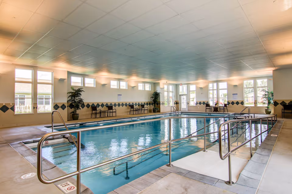 Indoor swimming pool with handrails and steps leading into the water, surrounded by a tiled deck. The room has large windows letting in natural light, potted plants, and chairs along the walls.
